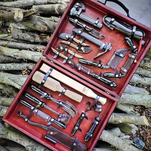 May include: A wooden box with a red interior, containing a collection of leather-wrapped tools and knives. The box is open and the tools are arranged neatly inside. The tools include a small axe, a saw, a knife, a chisel, and a hammer.