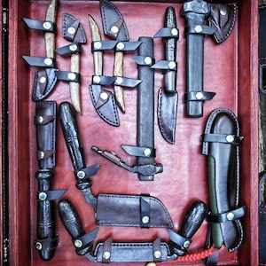 May include: A wooden box with a red interior containing a collection of leather-wrapped tools. The tools include a small axe, a knife, a saw, and several other tools. The tools are arranged in a neat and organized manner.