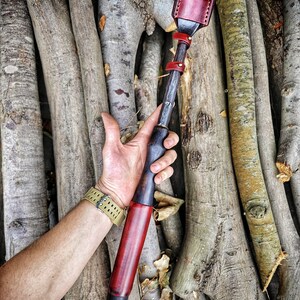 Peut inclure: Un outil en bois et en cuir avec une finition rouge et noire. L'outil est tenu dans une main sur un fond de bûches de bois empilées.
