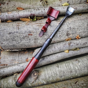 May include: A red leather sheath with a black handle and a metal blade. The blade is curved and has a sharp point. The sheath is attached to the blade with a metal loop. The blade is resting on a pile of logs.