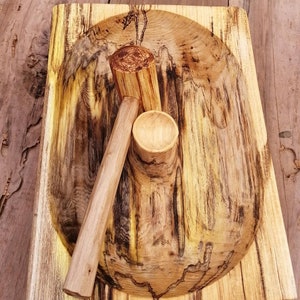 May include: A wooden mortar and pestle set. The mortar is a large, oval-shaped bowl with a dark brown and yellow wood grain. The pestle is a long, cylindrical piece of wood with a rounded head.