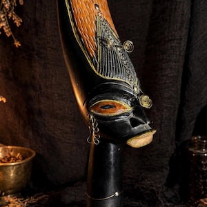 May include: A tall, black carved wooden sculpture of a stylized head with an elongated forehead, featuring textured brown wood grain, metallic wire accents, and a light-colored lip detail. A small brown paper tag is tied to the square base with twine.