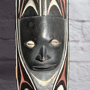 May include: A tall, vertical wooden carving featuring a central face with cowrie shell eyes. The surface is painted in black, white, and terracotta red with symmetrical, curved geometric patterns extending above and below the facial features.