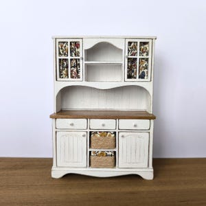 Puede incluir: Una alacena blanca en miniatura con encimera y cajones de madera. La sección superior tiene puertas con paneles de vidrio con inserciones de tela floral. Se ven dos cestas tejidas en el centro. Una pieza encantadora para una casa de muñecas o colección en miniatura.