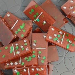 May include: A collection of brown dominoes with green leaf designs and silver dots. The dominoes are rectangular and have a shiny, transparent finish. The green leaves are arranged in a pattern on the surface of the dominoes.