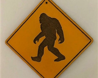 May include: Wooden Bigfoot crossing sign with a yellow background and a brown silhouette of Bigfoot walking. The sign is diamond-shaped with a small hole at the top for hanging. The edges of the sign are slightly darker.