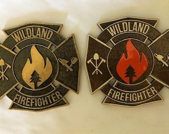 May include: Two Wildland Firefighter badges. One is gold with a gold flame and tree, the other is brown with a red flame and tree. Both have the words "Wildland Firefighter" and crossed tools.