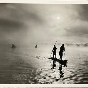 May include: A black and white photograph of three boats on a misty lake. Two people stand in the front boat, silhouetted against the water and sky. The sun is visible through the clouds.