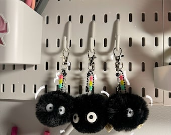 May include: Three black fuzzy keychain charms with white eyes hanging from a pegboard. Each charm has a rainbow beaded chain attached.
