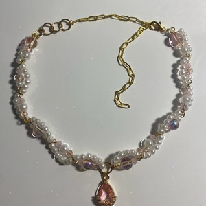 May include: A delicate gold chain necklace with a pink pear-shaped pendant. The necklace is made with white pearls and iridescent pink beads.