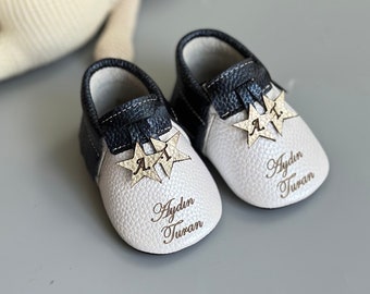May include: A pair of baby shoes featuring a white pebbled leather toe and a dark navy blue leather heel. Each shoe is adorned with two gold star-shaped charms engraved with the letters A.T. and the name Aydin Turan is printed in brown script.