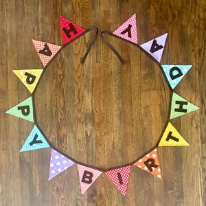 May include: A colorful "Happy Birthday" banner with triangular flags. Each flag features a different color and pattern, with letters spelling out the message. The banner is strung on a brown cord and laid on a wooden surface.