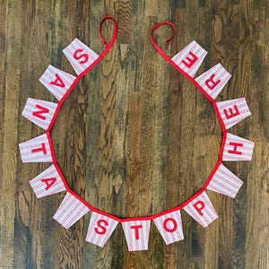 May include: A red and white striped fabric banner with the words "Santa Stop Here" in red letters. The banner is strung together with red ribbon and is hanging on a wooden floor.