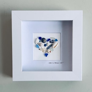 May include: A framed artwork featuring a heart-shaped arrangement of seashells, sea glass, and small starfish. The artwork is set against a white background within a white square frame. The piece is signed in the bottom right corner.