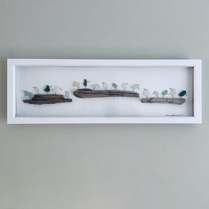 Può includere: Un'opera d'arte incorniciata che presenta una collezione di piccoli uccelli bianchi e verdi appollaiati su un pezzo di legno alla deriva. Gli uccelli sono fatti di vetro di mare e sono disposti in fila. L'opera d'arte è su uno sfondo bianco.