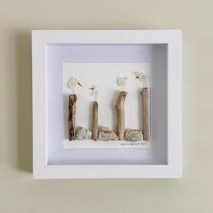 May include: Framed artwork featuring a seagull pebble art design. Four white seagulls are perched on driftwood sticks, with a white background and the text "Seagull Beach Art".