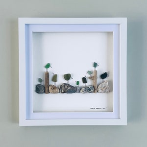 Puede incluir: Una obra de arte enmarcada que representa pájaros posados ​​en piedras y postes de madera. Los pájaros están hechos de vidrio verde y negro, con las piedras en tonos grises y marrones. La obra de arte está enmarcada en un marco blanco.