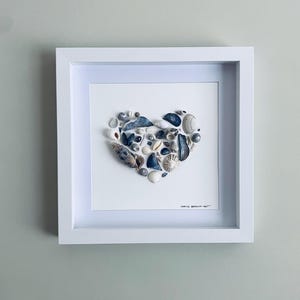 Può includere: Una cornice quadrata bianca a cassetta contenente una collezione di varie conchiglie e vetro marino disposti a forma di cuore su uno sfondo bianco. Il testo "DEB'S BEACH ART" è stampato in inchiostro nero nell'angolo in basso a destra.