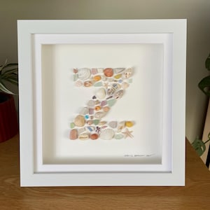 May include: A framed artwork featuring the letter "Z" crafted from various seashells and sea glass in shades of white, beige, orange, and blue. The artwork is set against a white background within a white frame. The text "1334'S BEACH ART" is visible.