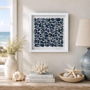 May include: A square white-framed wall art piece featuring a grid of dark blue mussel shells arranged on a white background. The shells are varied in size and shape, creating a textured, mosaic-like pattern within the frame.