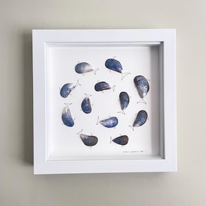 Peut inclure: Un cadre carré blanc contenant une œuvre d'art composée de dix coquilles de moules bleues disposées en cercle. Chaque coquille est fixée à un contour de queue de baleine dessiné à l'encre noire, créant l'apparence de baleines nageant. Le texte Libby's Beach Art est visible.