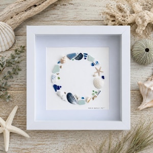 Op de afbeelding: Een vierkante witte lijst met een cirkelvormige schikking van diverse stukjes zeeglas, kleine schelpen en een zeester op een witte achtergrond. De tekst "ISAN'S BEACH ART" is in zwarte inkt gedrukt in de rechterbenedenhoek.