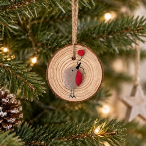 Può includere: Un ornamento circolare in fetta di legno con un pettirosso realizzato in sasso con il petto rosso e un piccolo cuore rosso attaccato a un filo. L'uccello ha zampe e becco disegnati a inchiostro nero. Un laccio in iuta serve per appenderlo.