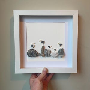 May include: A framed pebble art piece depicting seagulls on rocks and driftwood. The seagulls are made from white and grey pebbles, with black and yellow details. The artwork is set against a white backdrop and enclosed in a white square frame, suitable for coastal decor.