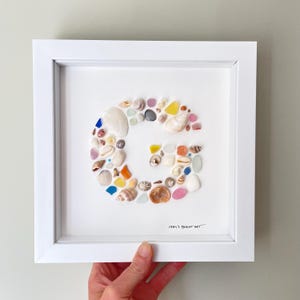 Puede incluir: Un marco cuadrado blanco que contiene una letra G formada por una disposición de varias conchas marinas pequeñas, vidrio marino y guijarros en tonos blanco, rosa, azul, amarillo y naranja. El texto 1994's BEACH ART aparece abajo a la derecha.