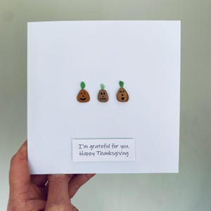 Puede incluir: Una tarjeta de felicitación blanca con tres pequeñas ilustraciones de calabazas marrones con tallos verdes y caras talladas. El texto "I'm grateful for you. Happy Thanksgiving" está impreso en un rectángulo blanco.