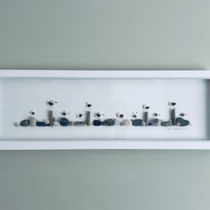 May include: A framed pebble art piece depicting a coastal scene. The artwork shows seagulls made of pebbles, perched on wooden posts and resting on pebbles. The frame is white, and the background is a light colour.