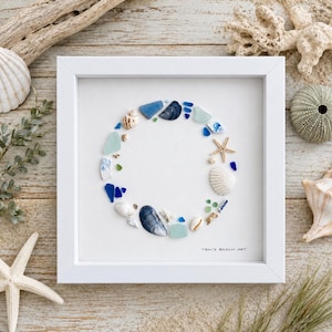 May include: A square white frame containing a circular arrangement of blue and white sea glass, small seashells, and a tiny starfish on a white background. The text TBM'S BEACH ART is printed in the bottom right corner of the white matting.