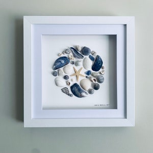 Puede incluir: Una obra de arte enmarcada en blanco con una disposición circular de conchas marinas y una estrella de mar en tonos azules, blancos y grises. La obra está firmada "ISONS BEACH ART".