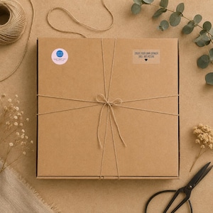 May include: A square brown cardboard box tied with thin twine. A circular sticker with blue wave icons and the text "Lucy's Beach Art" is on the top left. A rectangular label on the top right reads "CREATE YOUR OWN CORNISH SHELL ART PICTURE" with a heart.