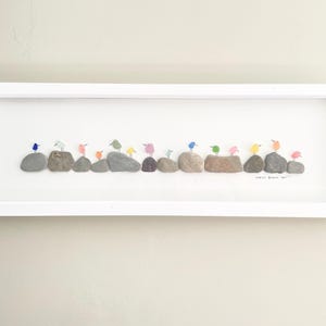 May include: A framed wall art piece featuring a row of small, colorful pebble birds perched on grey stones. The birds are painted in various pastel shades including blue, pink, yellow, and green. The artwork is set within a white rectangular frame.