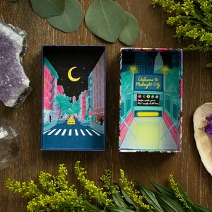 May include: A box with a colorful illustration of a city street at night with a crescent moon in the sky. The box is open and the inside is a bright yellow color with the text "Welcome to Midnight City".
