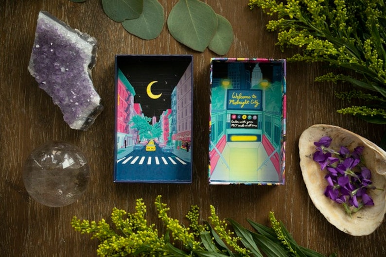 May include: A box with a colorful illustration of a city street at night with a crescent moon in the sky. The box is open and the inside is a bright yellow color with the text "Welcome to Midnight City".