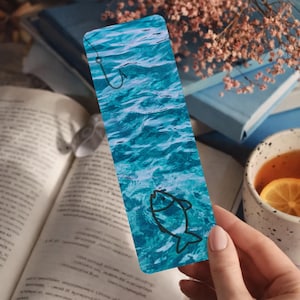 May include: A rectangular bookmark with a blue water design, a black fish outline, and a fishing hook. The bookmark is held over an open book, with a cup of tea and flowers in the background.