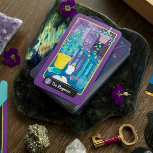 May include: A deck of tarot cards with a purple and gold design. The top card is The Magician, with a number 1 in the corner. The card features a colorful illustration of a potted plant, a candle, and a jar.