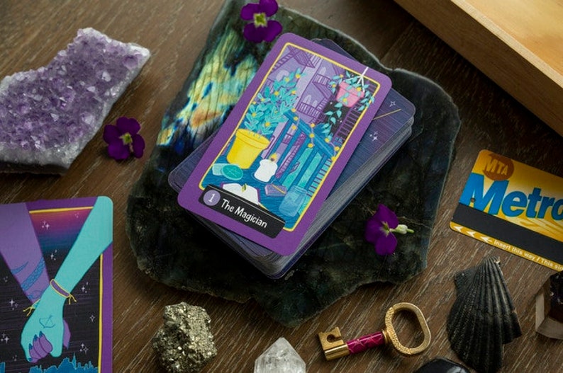 May include: A deck of tarot cards with a purple and gold design. The top card is The Magician, with a number 1 in the corner. The card features a colorful illustration of a potted plant, a candle, and a jar.
