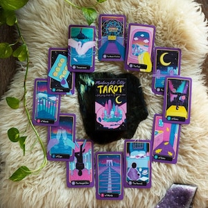 May include: A spread of tarot cards with a purple and gold design, featuring a city skyline and other illustrations. The cards are laid out on a white fluffy surface. The box for the deck is in the center of the spread and reads "Midnight City Tarot".