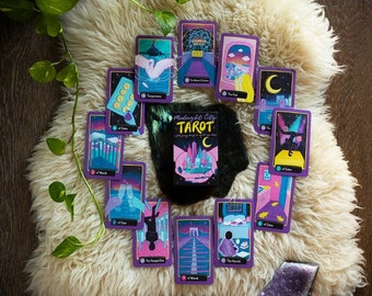 Könnte beinhalten: Eine Tarotkarten-Auslage mit einem violetten und goldenen Design, das eine Skyline der Stadt und andere Illustrationen zeigt. Die Karten liegen auf einer weißen, flauschigen Oberfläche. Die Schachtel für das Deck befindet sich in der Mitte der Auslage und trägt die Aufschrift "Midnight City Tarot".