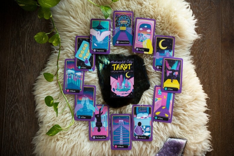 May include: A spread of tarot cards with a purple and gold design, featuring a city skyline and other illustrations. The cards are laid out on a white fluffy surface. The box for the deck is in the center of the spread and reads "Midnight City Tarot".