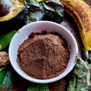 May include: A white ceramic bowl filled with a mound of dark brown powder, likely a spice blend. The bowl is sitting on a wooden surface with green leaves and a banana peel in the background.