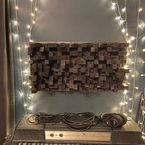 May include: A black sound-absorbing panel made of wood blocks, mounted on a wall with fairy lights. A black electric piano with a microphone and headphones sits in front of the panel.
