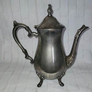 May include: A silver-colored teapot with a curved handle and spout. The teapot has a lid with a decorative finial and rests on three small feet. The design features ornate detailing on the handle, spout, and base, suggesting a vintage style.
