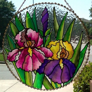 K&ouml;nnte beinhalten: Ein h&auml;ngendes Buntglasfenster zeigt drei lila, rosa und gelbe Irisbl&uuml;ten mit gr&uuml;nen Bl&auml;ttern. Das Fenster ist rund und hat einen goldenen Metallrahmen.