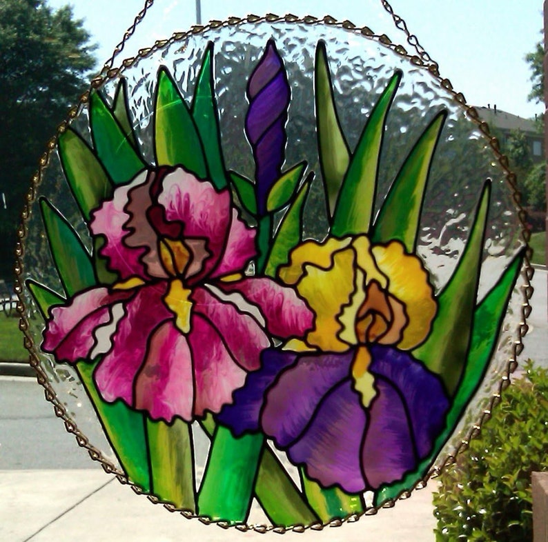 K&ouml;nnte beinhalten: Ein h&auml;ngendes Buntglasfenster zeigt drei lila, rosa und gelbe Irisbl&uuml;ten mit gr&uuml;nen Bl&auml;ttern. Das Fenster ist rund und hat einen goldenen Metallrahmen.