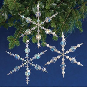 Könnte beinhalten: Zwei dekorative Schneeflocken-Ornamente mit schillernden Kristallen und silberfarbenen Metallakzenten. Eine Schneeflocke hat klare und blaue Kristalle, die andere weiße perlenartige Perlen. Grüne Kiefernzweige sind im Hintergrund.