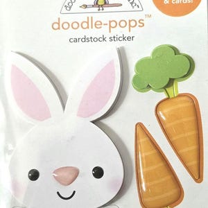 May include: Doodlebug Design Inc. "doodle-pops" cardstock sticker set featuring a white bunny with pink ears and paws, and two orange carrots with green tops. The sticker set is designed for scrapbooks and cards.